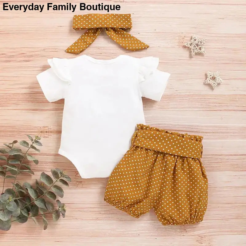 White baby bodysuit with mustard yellow polka dot shorts and matching bow hair accessory.