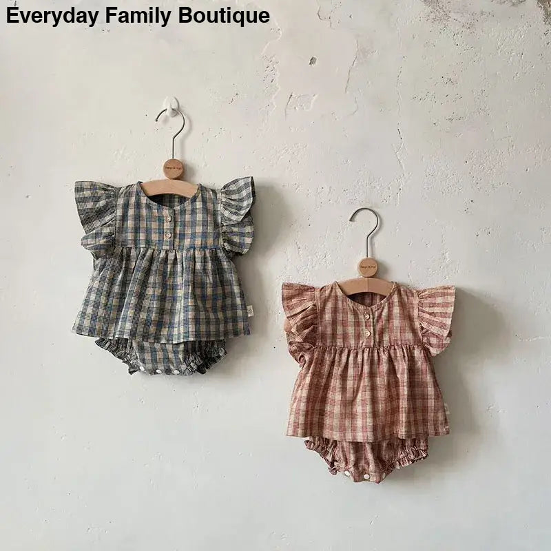 Two plaid baby outfits with ruffled sleeves and matching bloomers hanging on wooden hangers.