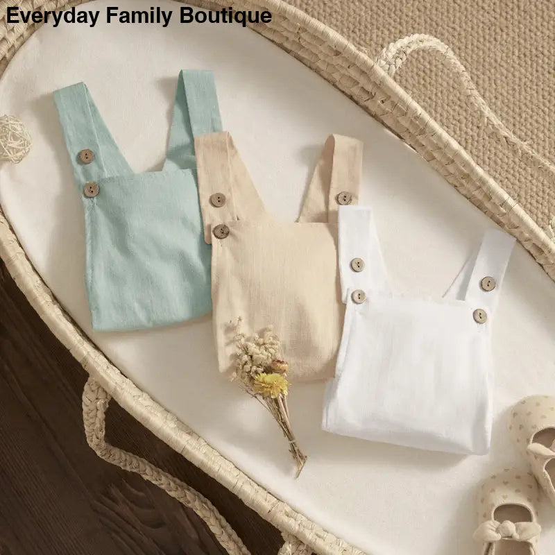 Three miniature linen overalls in mint green, beige, and white colors with decorative buttons.