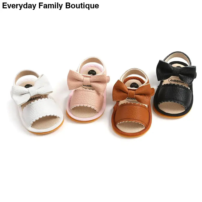 Four baby sandals with bows in different colors: white, pink, tan, and black.