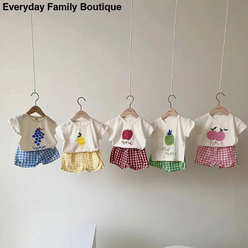 Five children’s outfits with white t-shirts and gingham shorts in different colors hanging on wooden hangers.