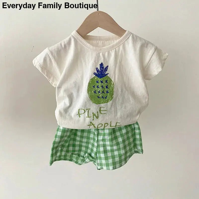 White t-shirt with a pineapple design paired with green gingham shorts.