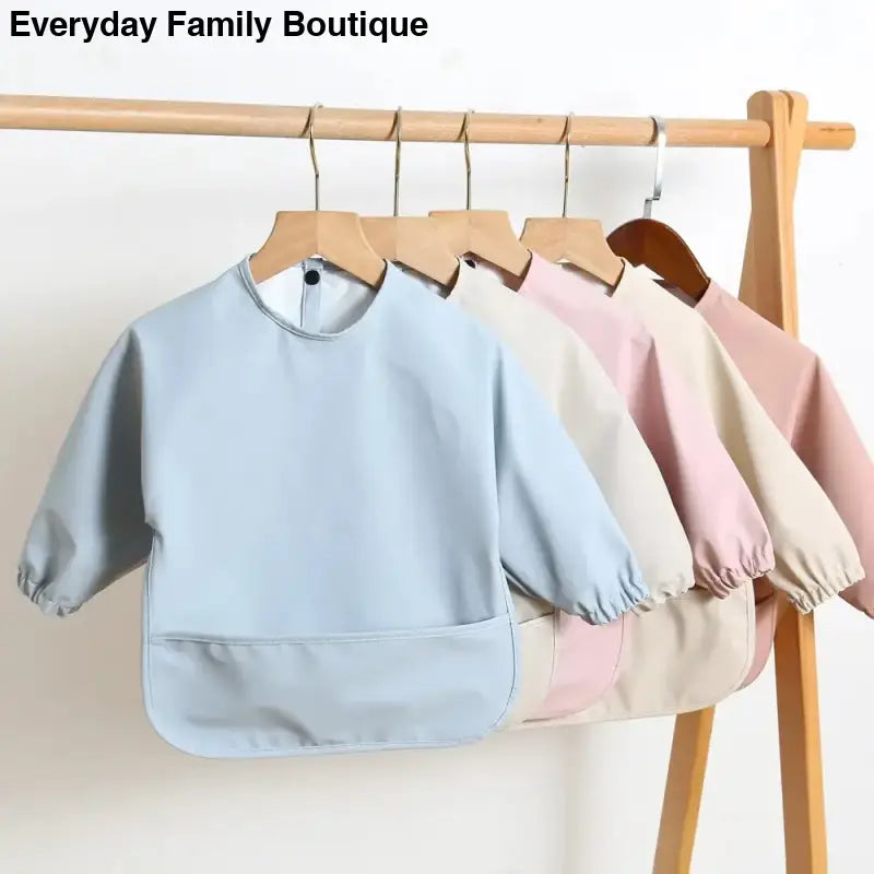 Long-sleeved waterproof bibs in pastel colors hanging on wooden hangers.