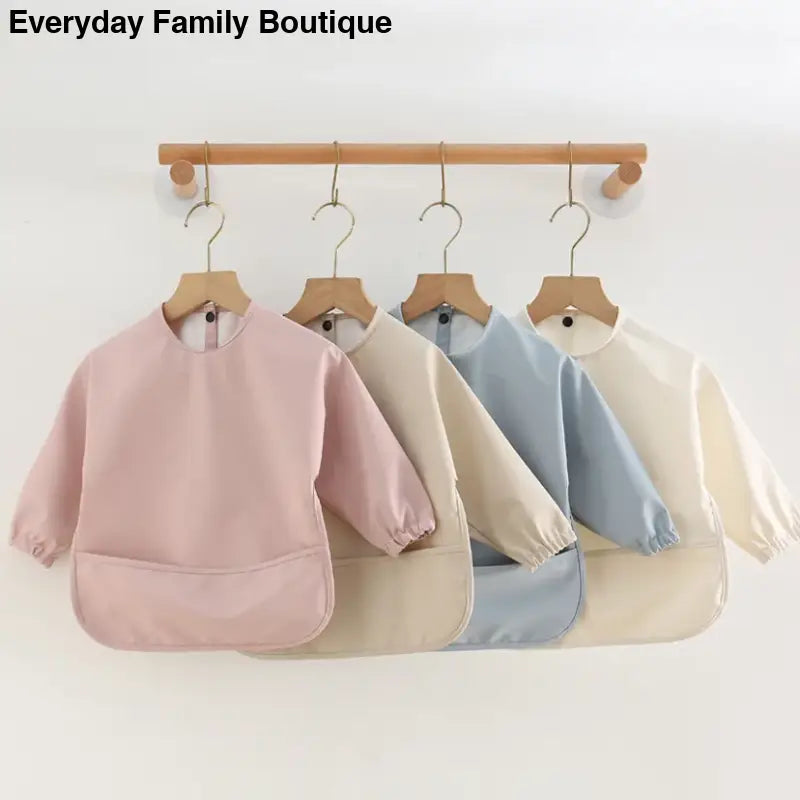 Long-sleeved waterproof bibs in pastel pink, beige, blue and cream colors hanging on wooden hangers.